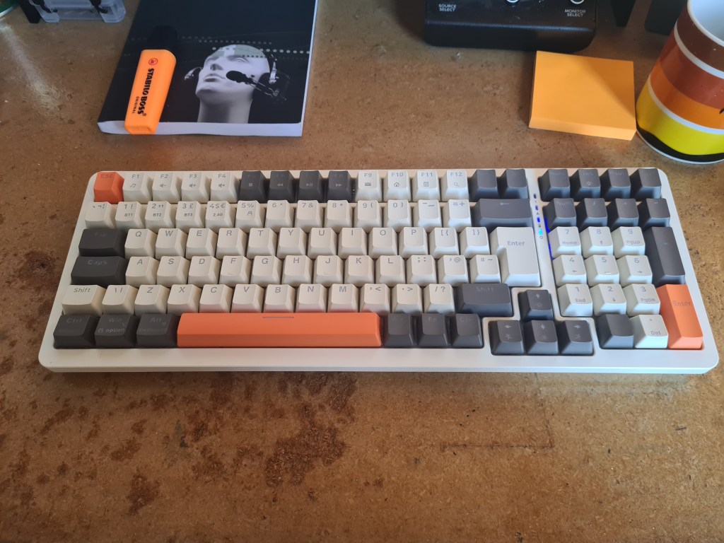 A wireless mechanical computer keyboard on a wooden desk. Also in shot: post-it notes, a coffee mug, a highlighter pen, and a copy of the book 'Listening through the noise: the aesthetics of experimental electronic music', by Joanna Demers.